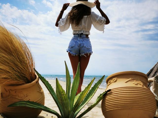 PassionX stands with her back to the camera, wearing a white blouse and frayed denim shorts, holding a white hat, with her ass prominently visible, by the ocean.