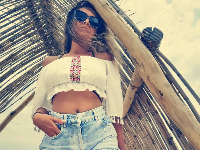 PassionX with sun-kissed skin, wearing a white off-shoulder crop top with pink embroidery, denim jeans, and dark sunglasses, standing under a wooden, thatched roof structure.