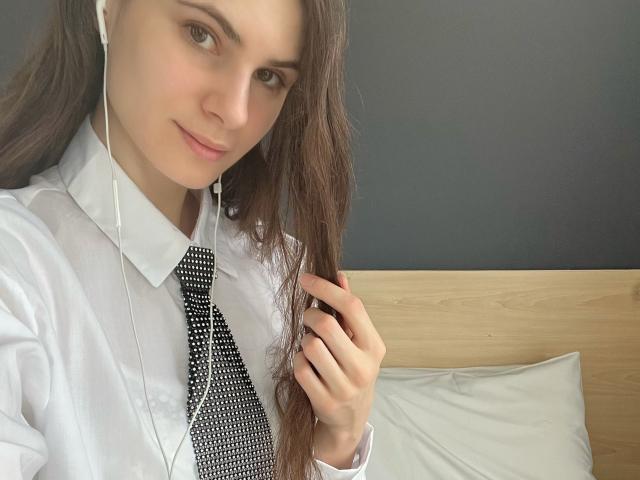 BatSama con lunghi capelli castani, camicia bianca, cravatta a pois bianchi e neri, auricolari, che si tiene i capelli, sorride dolcemente, pelle chiara, trucco leggero, testiera di legno, cuscino bianco, parete grigia..