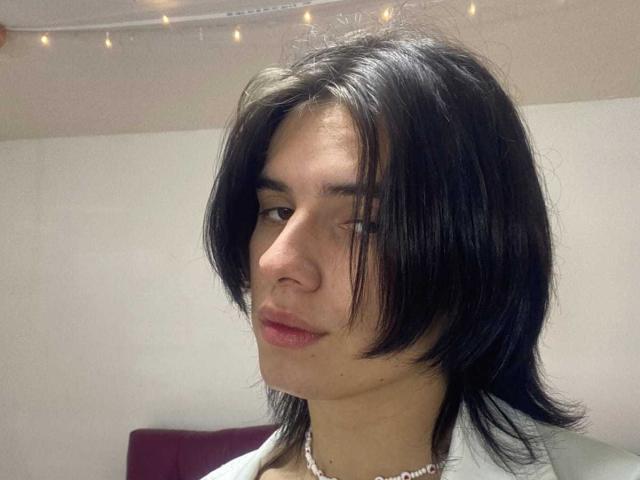 Brooke69 a une coupe de cheveux noire mi-longue, elle porte une chemise blanche avec un collier de perles, et elle a une expression sérieuse, avec un maquillage discret et des lèvres d'un rose clair..