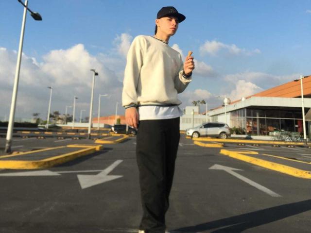 Brooke69 steht auf einem Parkplatz, trägt eine schwarze Mütze, einen weißen Pullover und eine schwarze Hose und zeigt mit dem Finger eine Geste, mit ernstem Gesichtsausdruck, unter blauem Himmel..