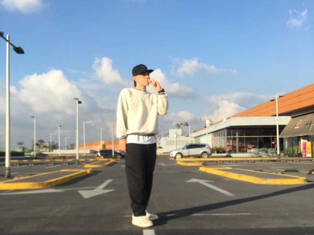 Brooke69 está em um estacionamento, usando um boné preto, uma jaqueta de moletom branca, calças pretas e tênis brancos, com um telefone no ouvido. Céu azul com nuvens..