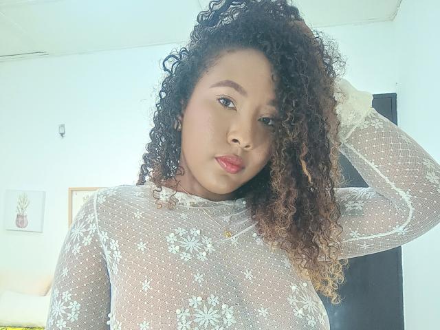 CaroldelaTorre with curly brown hair, wearing a sheer white lace top, pouting with pink lips, hand in hair, looking at camera, small gold necklace, soft natural light.