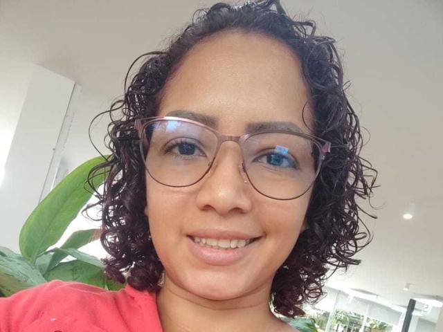 AnaferHot, com cabelos castanho-escuros e cacheados, usando óculos rosa e uma camisa rosa, sorrindo mostrando os dentes, tem olhos azuis e um tom de pele médio..