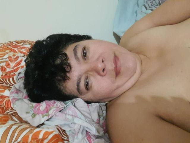 Angellovelly is lying on a bed, shirtless, with curly black hair, brown eyes, and a slight smile, showing a chubby chest and a light skin tone.