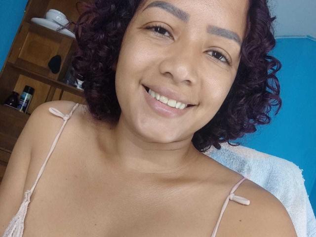 AnaferHot, com cabelo roxo encaracolado, sorrindo, vestindo uma blusa rosa com alças, mostrando seus seios bonitos e nádegas macias em um selfie. Seus olhos castanhos e expressão relaxada são visíveis..