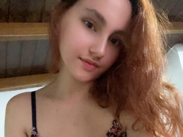 EstefaniaRamos, avec ses cheveux ondulés et châtain-roussis, sa peau claire et un maquillage discret, porte un soutien-gorge noir à motifs floraux et sourit doucement à l'appareil photo..