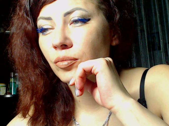 JuliaIce with curly brown hair, blue winged eyeliner, and glossy lips, resting her chin on her hand, wearing a black strap, and a silver necklace.