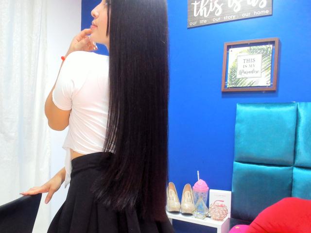 SarahGood with long black hair, white crop top, and black skirt, standing in room, biting finger, blue wall, turquoise headboard, and pink cupcake decoration in background.