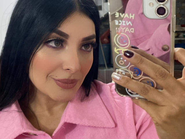 ArianaCortes, avec des cheveux noirs, du rouge à lèvres rose et du fard à paupières violet, portant une chemise rose, tenant un téléphone avec des motifs colorés, regardant un selfie dans un miroir..
