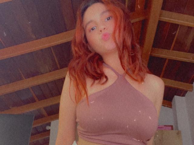 EstefaniaRamos, una donna attraente con i capelli rossi, labbra piene, che indossa un top rosa con spalline, mostrando il suo seno generoso, in una stanza con soffitto in legno. Sta guardando la fotocamera..