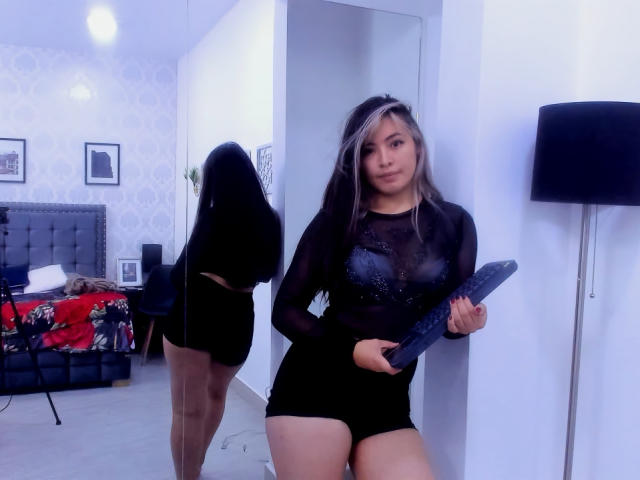 SallyDoll mit langem, schwarzem und blondem Haar, die ein durchsichtiges schwarzes Oberteil und Shorts trägt, hält einen blauen Dildo und steht vor einem Spiegel, der ihren Hintern reflektiert..