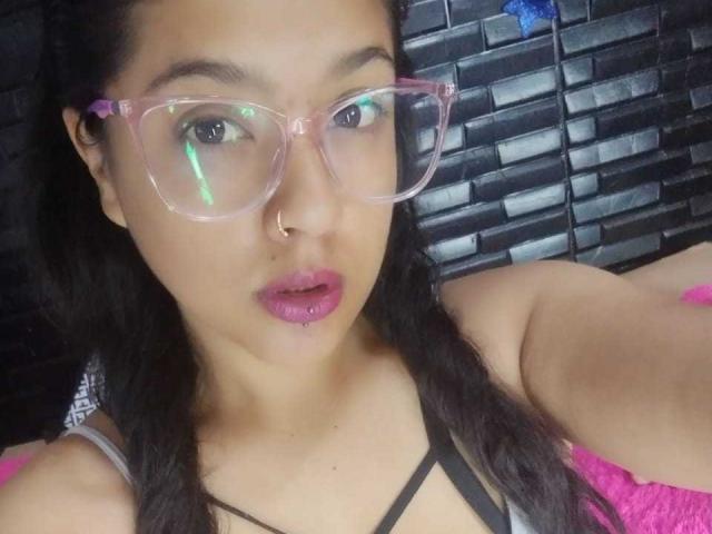 Brunette, Lia, avec des lunettes à monture rose, un anneau de nez et du rouge à lèvres rose, porte un haut noir à bretelles, on peut voir les bretelles roses de son soutien-gorge, et ses cheveux foncés sont lâchés..
