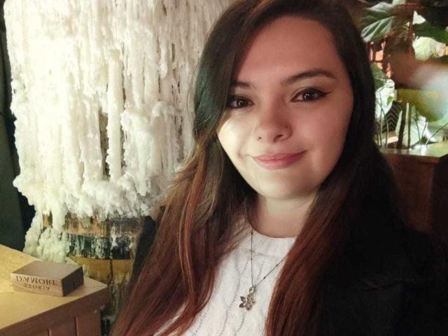 KatherinLatin has long brown hair, a cute smile, and is wearing a black jacket over a white shirt with a necklace. Her eyeliner is winged and she's looking at the camera.