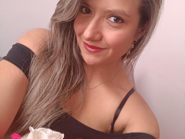 ValeriaHunter69 with long, grayish-brown hair, smiling, wearing an off-shoulder black top, pink lipstick, and gold earrings, showing her chest with a black strap.