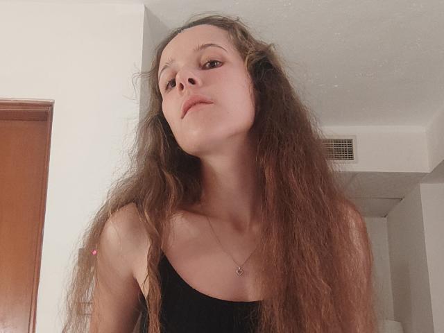 JollyKiss69, avec de longs cheveux bruns en désordre, portant un débardeur noir, regardant vers le haut avec un léger sourire, un collier avec un petit pendentif en forme de cœur, peau claire, maquillage minimal..