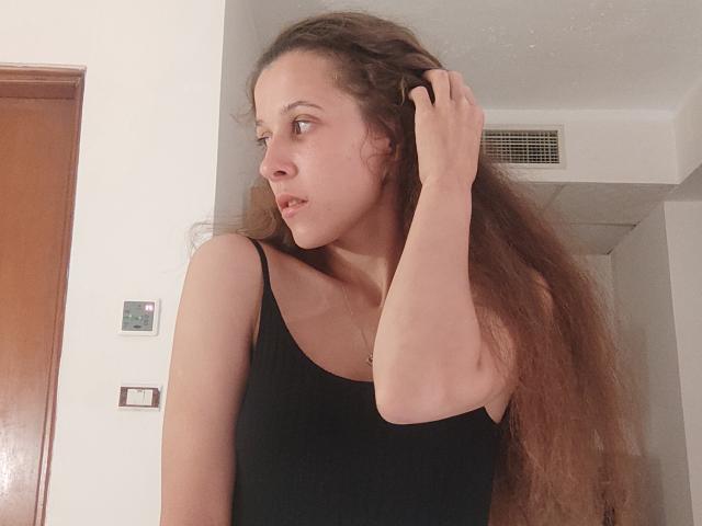 JollyKiss69 com cabelos castanhos longos e ondulados, vestindo uma regata preta, com a mão no cabelo, perfil lateral, olhando para a esquerda, boca ligeiramente aberta, pele clara, ambiente interno casual..