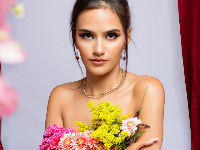 AmelieLautrec with a serious expression, holding colorful flowers, bare shoulders, gold necklace, and red earrings, her tits slightly visible, brown eyes, and dark hair pulled back.