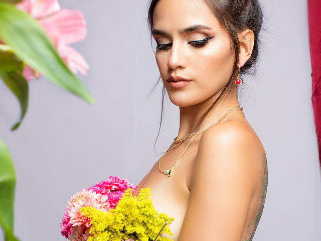Amelie Lautrec, com cabelo escuro, delineador rosa, brincos vermelhos, colar dourado, segurando flores coloridas, cobrindo seus seios nus, uma flor rosa em primeiro plano, flores amarelas, perfil lateral, sorriso discreto..
