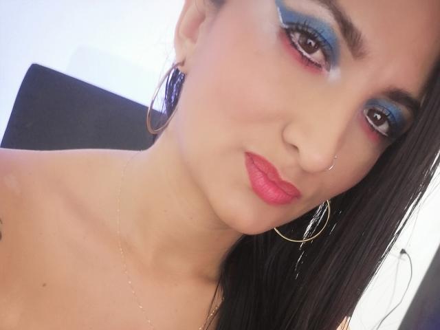 Feetabbe with blue eye makeup, pink lipstick, hoop earrings, nose ring, and long black hair, looking at the camera. Her tits are partially visible. She's wearing a thin necklace.
