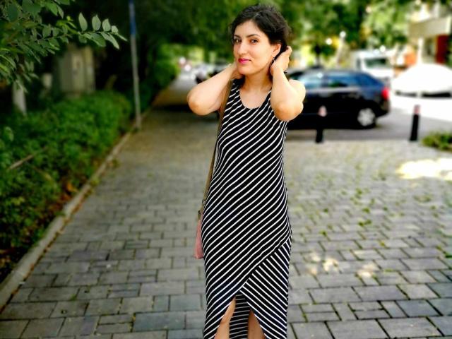 AddictedToUTS stands on a brick sidewalk, wearing a black and white striped dress, with hands in her curly dark hair, showing a slight smile, in a sunny urban setting.