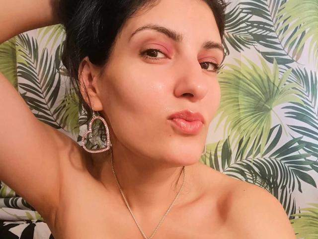 AddictedToUTS with pouty lips, pink eyeshadow, shiny skin, dark hair, pink earrings, gold necklace, arm raised, bare shoulders, green leaf pattern background.