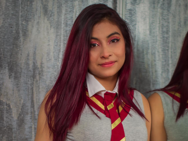 Samantha Smith, met lang paars haar, roze eyeliner en een rood-geel gestreept dasje over een wit shirt, glimlacht zachtjes en is weerspiegeld in een spiegel. Ze draagt een grijze mouwloze top en heeft een gemiddelde borstmaat..