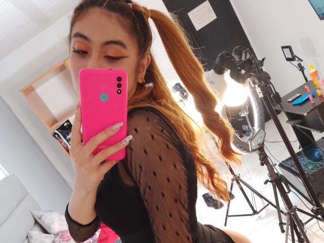 Samantha Smith, con largas trenzas marrones, vistiendo una blusa negra transparente con lunares, sosteniendo un teléfono rosa, tomando una selfie en el espejo, mostrando su trasero, en una habitación muy iluminada..
