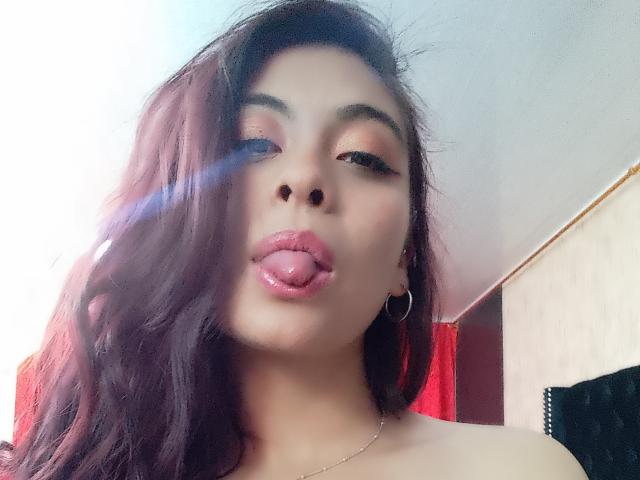 SamanthaaSmith with dark, wavy hair, sticking out her glossy pink tongue, wearing hoop earrings, and a thin necklace, in a selfie with a lens flare.