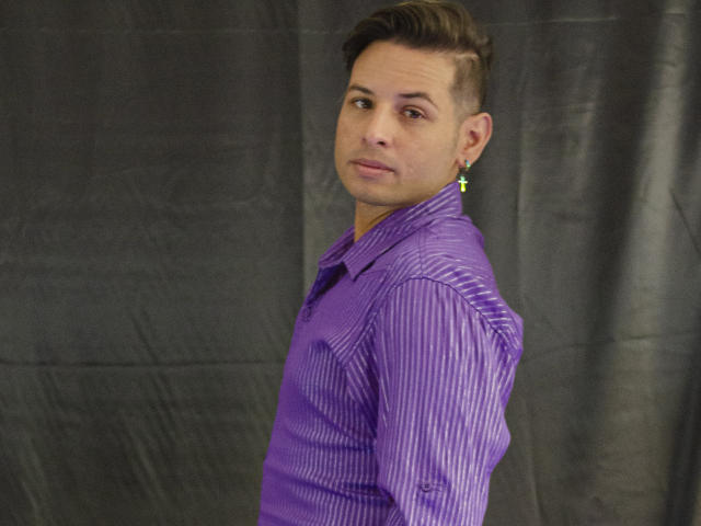 JeanSmithHot, con cabello corto y castaño, vistiendo una camisa morada a rayas y un arete verde, se gira de lado, mirando por encima del hombro, mostrando una leve sonrisa..