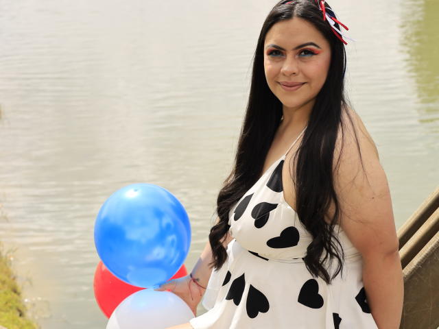 ZoeyJones with long black hair, wearing a white dress with black heart patterns, holding blue, red, and white balloons, standing by a water body, smiling.