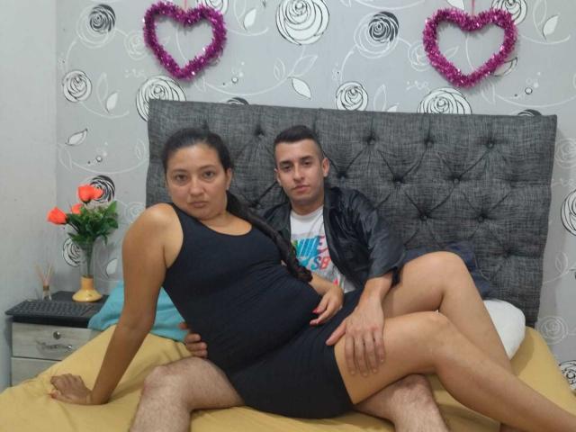 MaxAndViolet, een zwangere vrouw met een zwart tanktop en zwarte korte broek, zit op een bed met haar billen en vagina bloot, terwijl een man met een leren jas erachter zit..