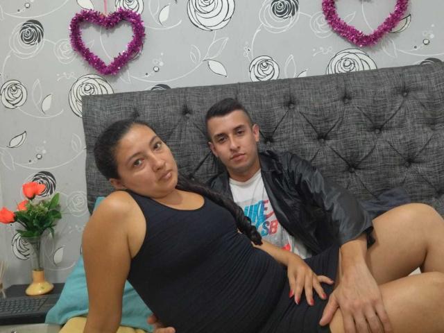 MaxAndViolet, uma mulher latina com cabelo preto, vestindo uma camiseta e um vestido preto, está sentada na cama, com as nádegas apoiadas, ao lado de um homem latino vestindo uma camisa..