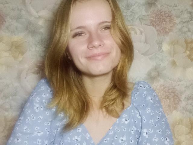 MargaritaKis with blonde hair, light skin, smiling softly, wearing a blue floral dress, showing her cute face and small tits, standing against a floral-patterned wall.