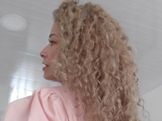 Dalila, con cabello rubio rizado, camisa rosa, vista de perfil, sonriendo, nariz visible, techo blanco, luz suave, ambiente relajado. Los senos no son visibles, el enfoque está en el rostro y el cabello..
