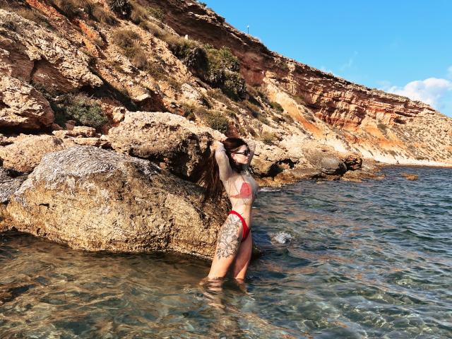 ValeriMair debout dans une eau claire, portant un bikini rouge, tenant un rocher, avec des tatouages sur la cuisse gauche et le bras droit, devant une falaise rocheuse, par une journée ensoleillée..