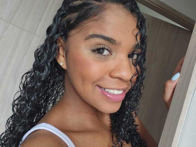 LisaMendez ha i capelli neri e ricci, la pelle marrone ed è sorridente con le labbra rosa, indossa una maglietta bianca, mettendo in risalto il suo viso carino e l'unghia blu. Sta tenendo una porta..