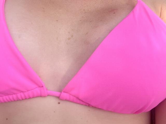 Il top rosa del bikini di NancyChaude, che mostra i suoi piccoli seni, leggermente macchiati e abbronzati, con una leggera linea di abbronzatura e un piccolo neo appena sotto il seno sinistro..