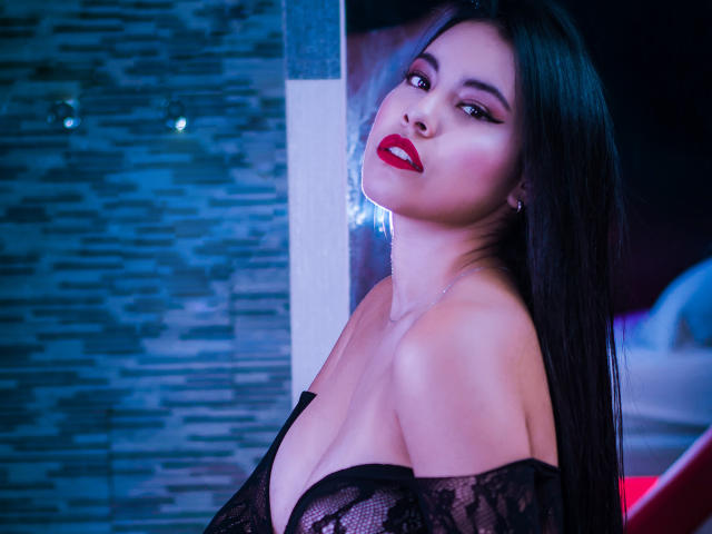 PaolaYKatty, con lunghi capelli neri, rossetto rosso e una top nera in pizzo con spalline scoperte, che mostra il seno, guarda seducentemente la fotocamera, in una stanza illuminata di blu..