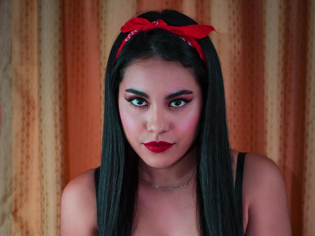 PaolaYKatty with long black hair, red bandana, and red lipstick, staring directly at the camera, showing off her puffy eyeliner and cute face, with her tits barely visible.