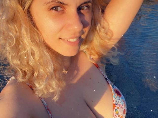 LucilleXSugar met krullend blond haar, lachend naar de camera, gekleed in een bikini met bloemenprint, waarbij haar borsten zichtbaar zijn in het zonlicht, met armen omhoog..