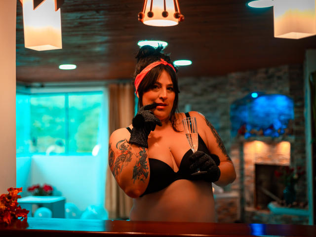 Isabella Roshe con un sujetador negro, guantes negros, una diadema roja, un brazo tatuado, señalando, sosteniendo la tira del sujetador, techo de madera, luces colgantes, iluminación azul y cálida, fondo ligeramente borroso..