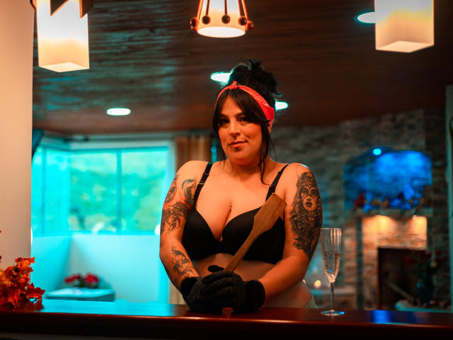 IsabellaRoshe with black bra, tattoos on arms, red headband, black gloves, holding wooden spoon, standing at bar, glass of champagne, wood ceiling, warm lights, slight smile, medium tits.