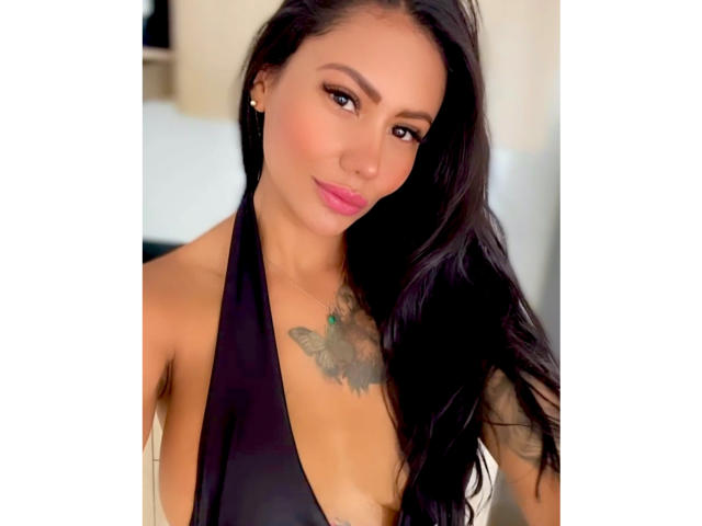 AlessandraMalory with long black hair, brown eyes, and pink lipstick, wearing a black halter top, showing a tattoo on her chest, and small earrings. Boobs visible, medium-sized.