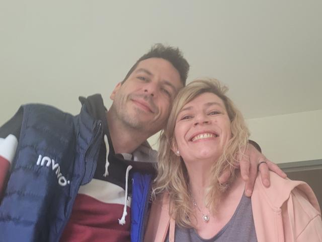 OscarBella mit kurzen, dunklen Haaren, leichtem Bart und einer blauen, gepolsterten Weste, steht neben OscarBella mit blonden Haaren, einem grauen Oberteil und einer pinken Jacke, beide lächeln..