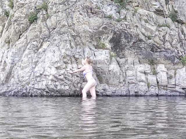 SweetAleksaX, blond, draagt een paars bikini, staat in ondiep water, handen tegen een rotswand, billen licht omhoog, water reflecteert haar lichaam, rotswand op de achtergrond..