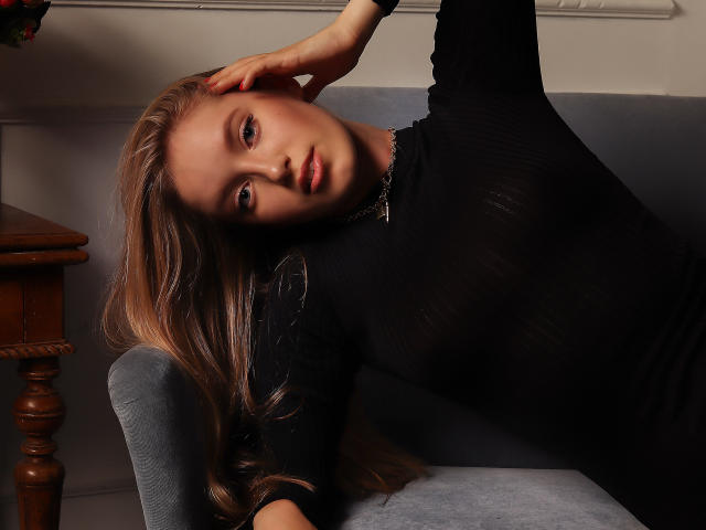 EvaPink, com cabelos loiros longos, usando uma blusa preta transparente, deitada em um sofá cinza, com a cabeça inclinada, um braço apoiado, olhando para a câmera, maquiagem discreta, colar, mesa de cabeceira de madeira, iluminação suave..