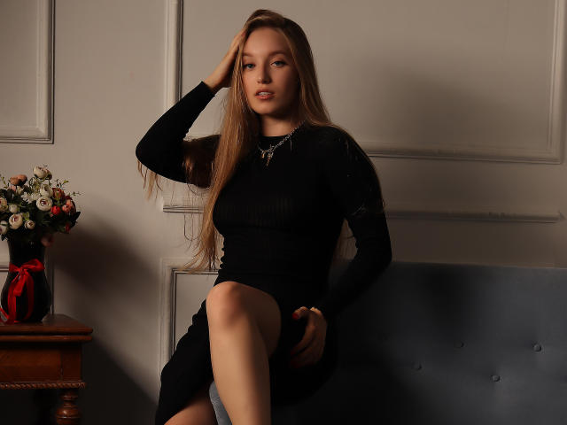 EvaPink, con lunghi capelli castani, vestito nero e collana, seduta con una gamba sollevata, mano tra i capelli, che guarda la fotocamera, vaso di fiori su un tavolo di legno, seduta imbottita blu..