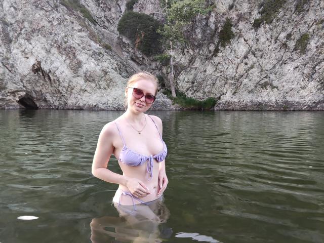 SweetAleksaX de pie en el agua, con un top de bikini color lila, sus pequeños senos visibles, con las manos sobre la barriga, gafas de sol puestas, un acantilado rocoso al fondo, follaje verde..