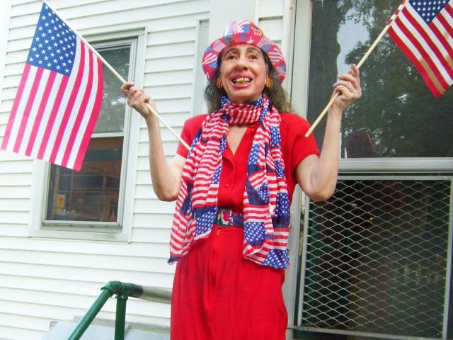 ElenahHottie, met een rode jurk, een gestreepte sjaal, een hoed en Amerikaanse vlaggen, staat lachend, vlaggen vasthoudend, haar borsten onthullend en een riem dragend, voor een wit gebouw..